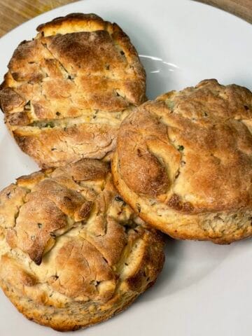 Three expertly baked golden-brown biscuits are stacked on top of each other on a white plate on a wooden cutting board next to a lit candle on a white countertop