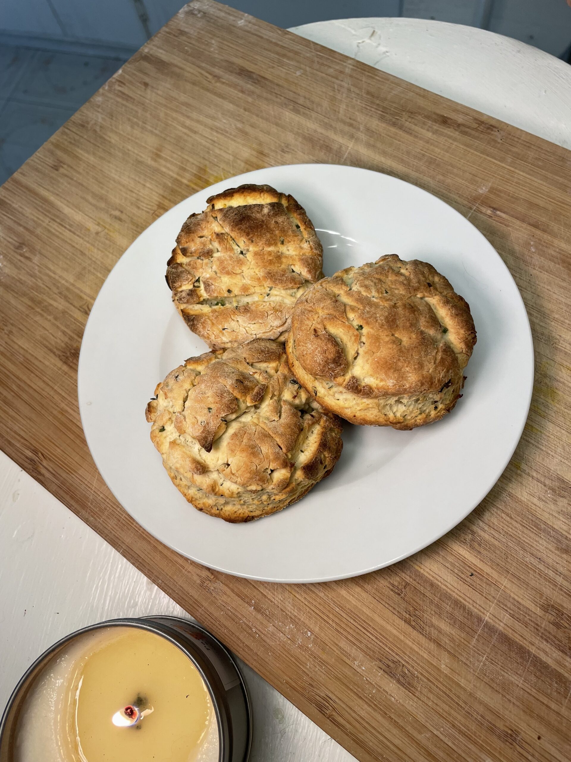 Three expertly baked golden-brown biscuits are stacked on top of each other on a white plate on a wooden cutting board next to a lit candle on a white countertop