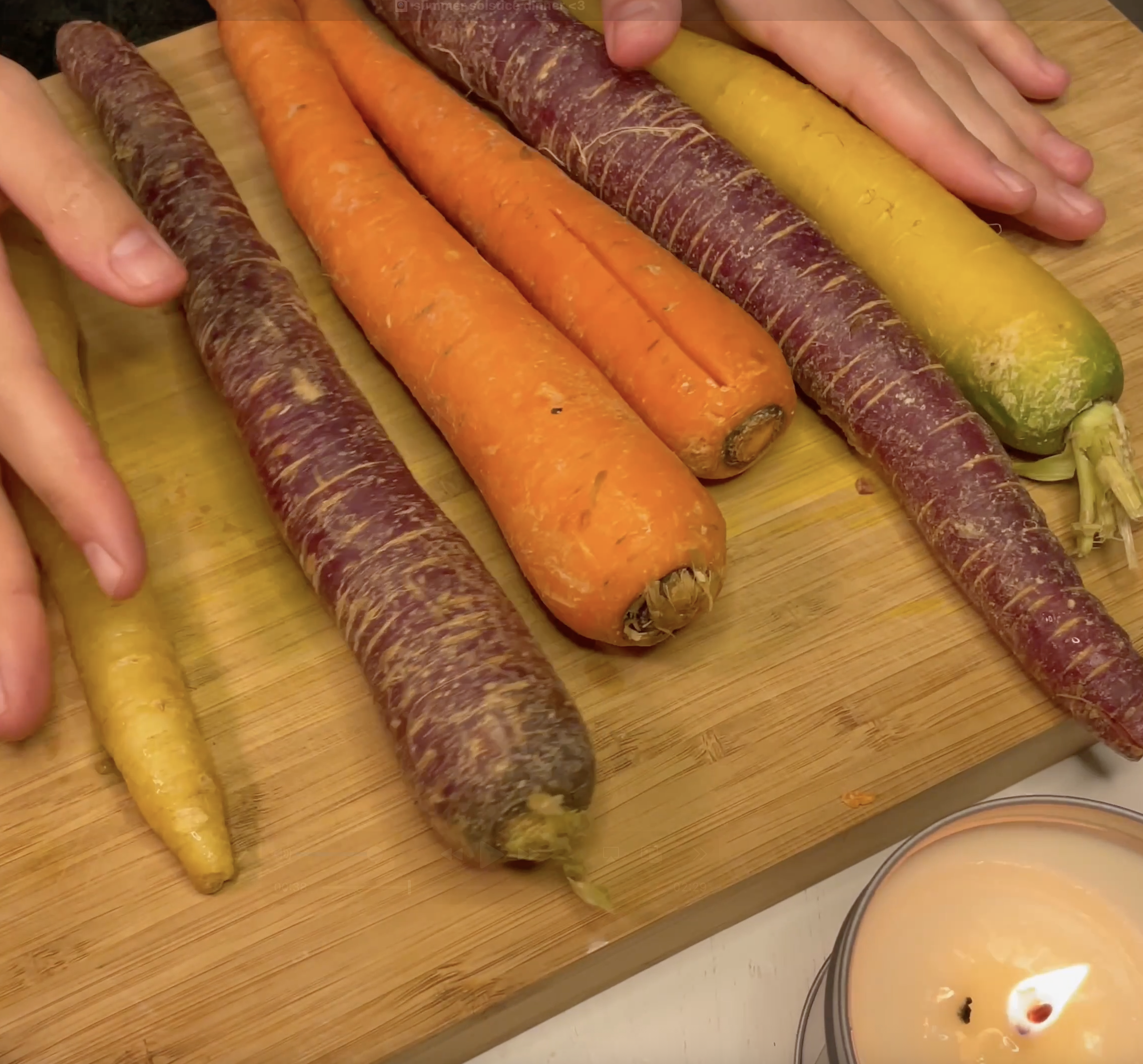 Rustic carrots colored in vibrant purple, yellow and classic orange are laid out on a wood cutting board. On the top of the picture, there is a hand keeping the carrots in place. 