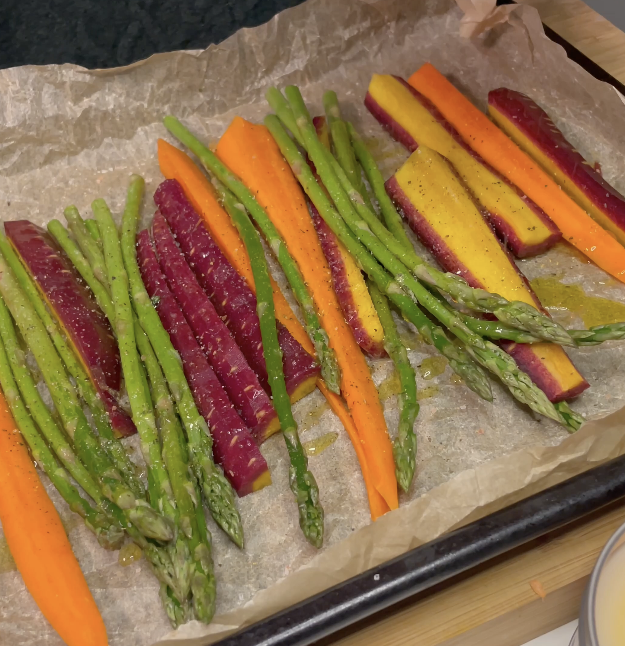 Brightly colored carrots, purple, yellow and orange, and vibrant green asparagus are expertly seasoned on a black baking sheet on brown parchment paper. 