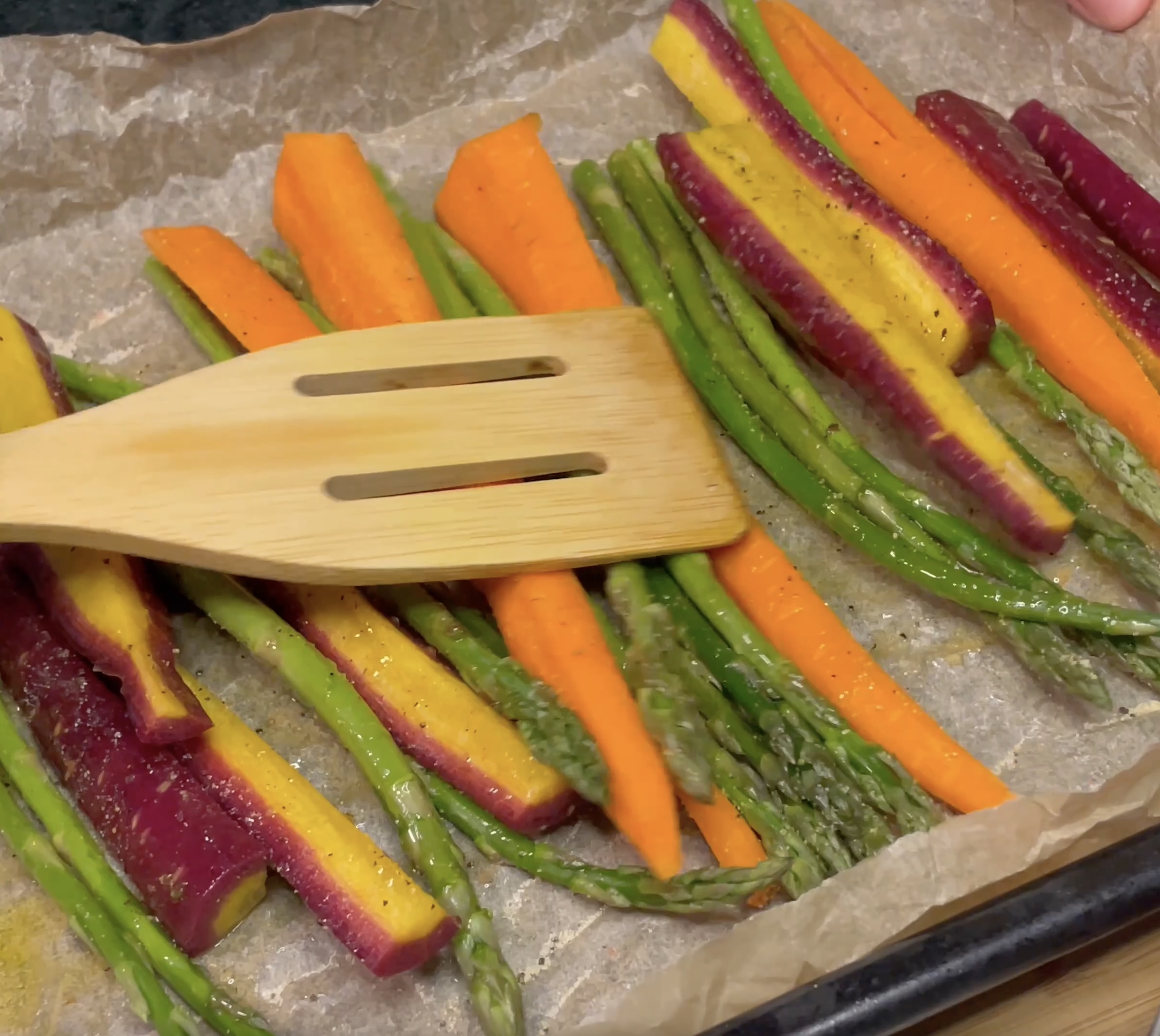 Brightly colored carrots, purple, yellow and orange, and vibrant green asparagus are expertly seasoned on a black baking sheet on brown parchment paper after being cooked in the oven, flipping with a rustic, light brown wooden spatula. 