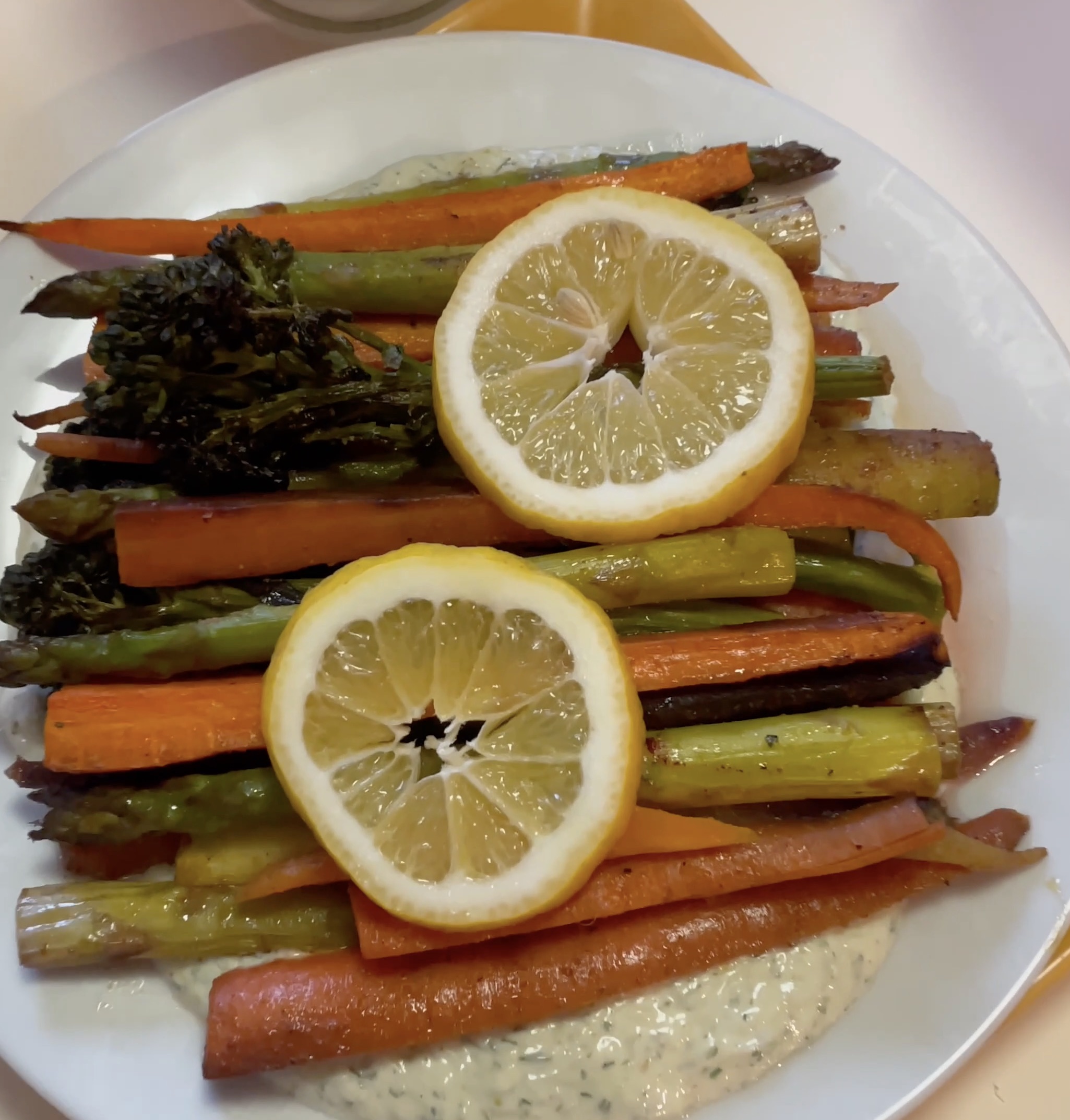 On a shiny, white plate lays a bed of tzatziki underneath perfectly seasoned and cooked orange, yellow, and purple carrots and green asparagus. This dish is garnished with bright yellow lemon slices. 