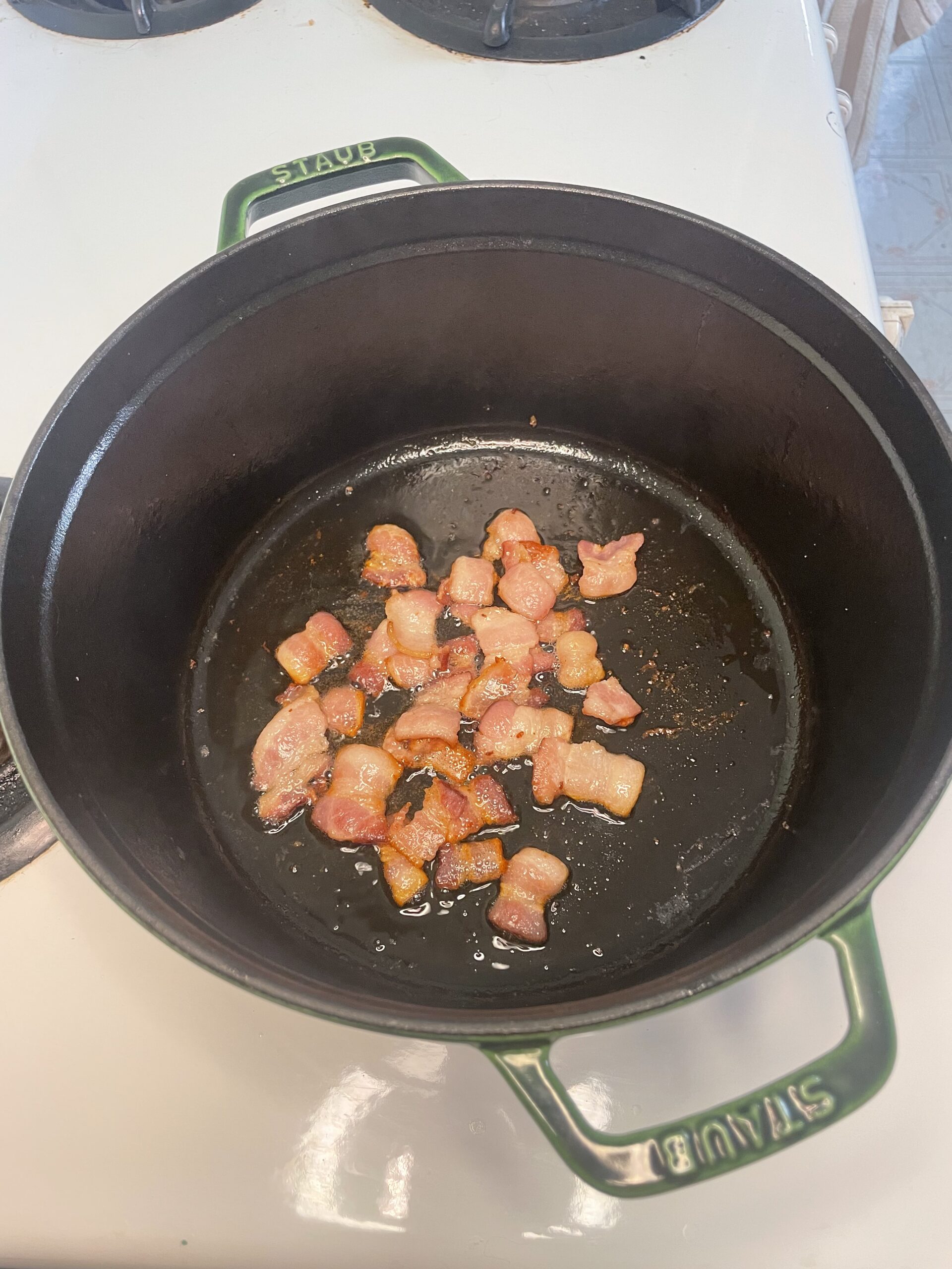 On a white stove, cooking in a green Staub pot is caramelized bacon rendering bacon fat into the pot.