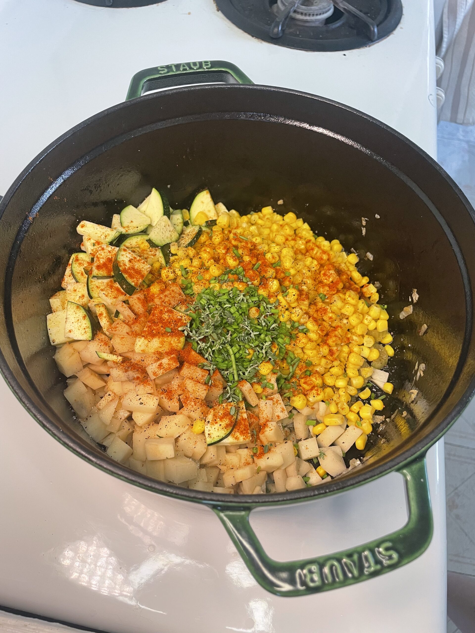 In a green Staub pot, on top of the cooked bacon there is cubed potato, cut zucchini, golden corn, green herbs in the middle, as well as red paprika sprinkled on the top.