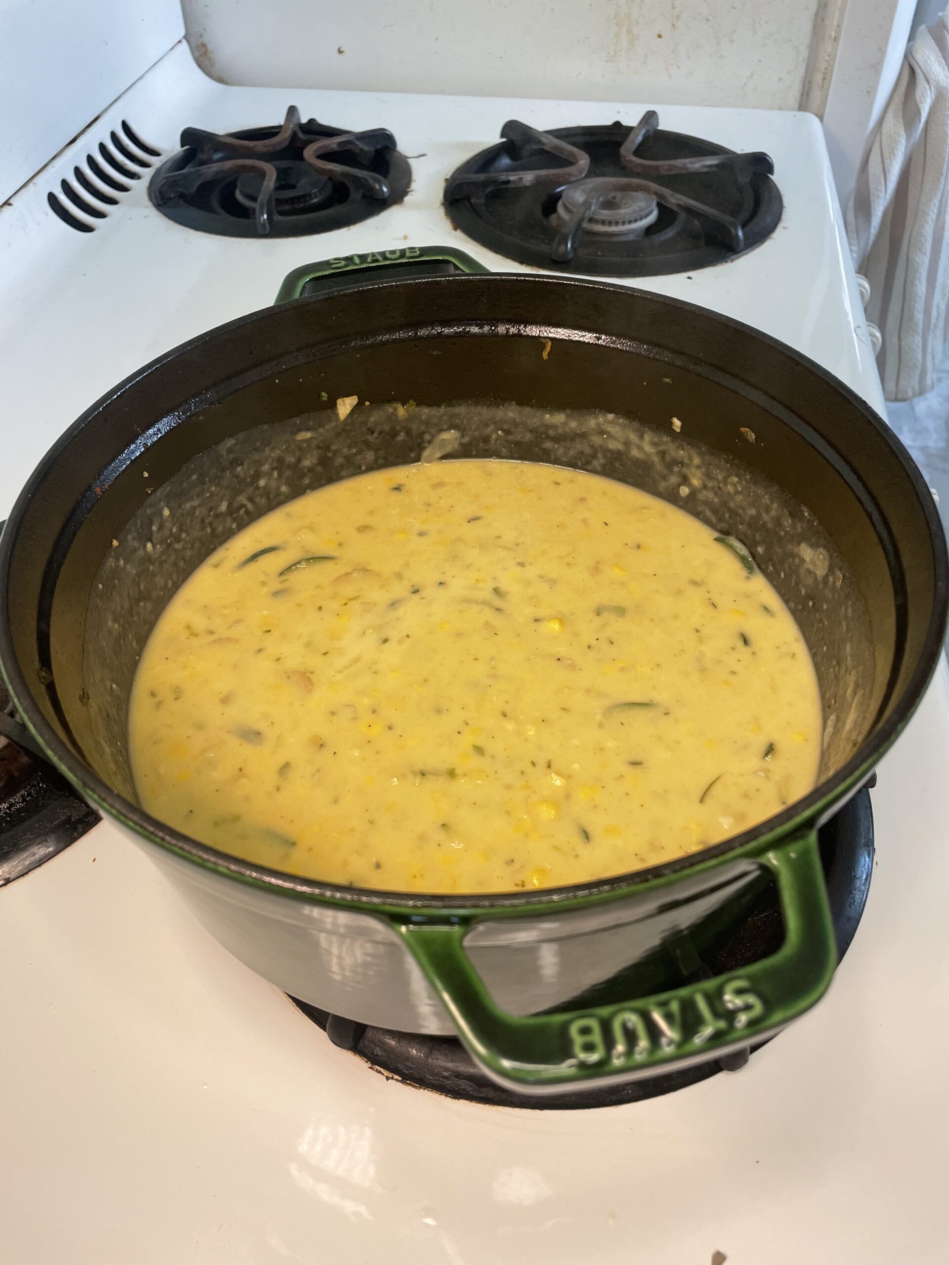 On a white stove in a green Staub pot, yellow soup is cooking with green and yellow vegetables throughout.
