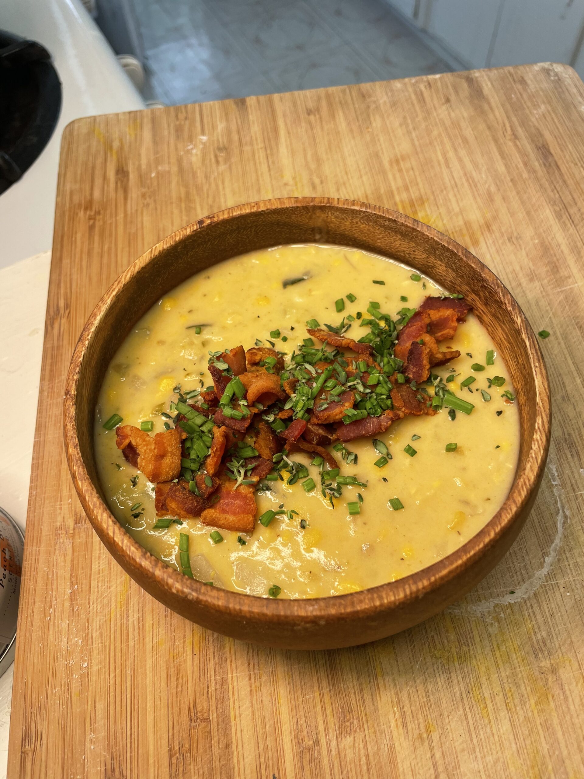 On a wood cutting board, a wooden bowl of yellow soup filled with vegetables is garnished with cut, cooked bacon and green onion.