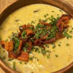 On a wood cutting board, a wooden bowl of yellow soup filled with vegetables is garnished with cut, cooked bacon and green onion.