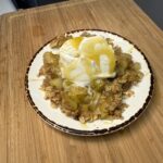 The image shows a plated serving of Caramel & Chamomile Pear Crumble placed on a wooden cutting board. The crumble consists of tender, golden pear pieces baked with an oat-based topping. On top sits a generous scoop of vanilla ice cream, melting slightly, and drizzled with a glossy caramel sauce that cascades over the dessert and onto the plate. The rustic plating and warm tones give it a cozy, homemade feel.