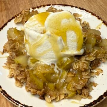 The image shows a plated serving of Caramel & Chamomile Pear Crumble placed on a wooden cutting board. The crumble consists of tender, golden pear pieces baked with an oat-based topping. On top sits a generous scoop of vanilla ice cream, melting slightly, and drizzled with a glossy caramel sauce that cascades over the dessert and onto the plate. The rustic plating and warm tones give it a cozy, homemade feel.