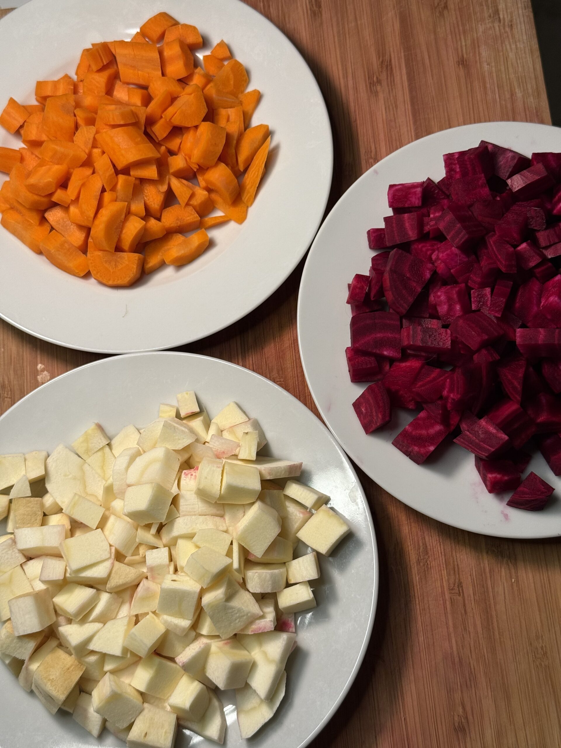 On a wooden cutting board, there are three white plates, one with cubed orange carrots, cubed purple beets, and cubed parsnips. 