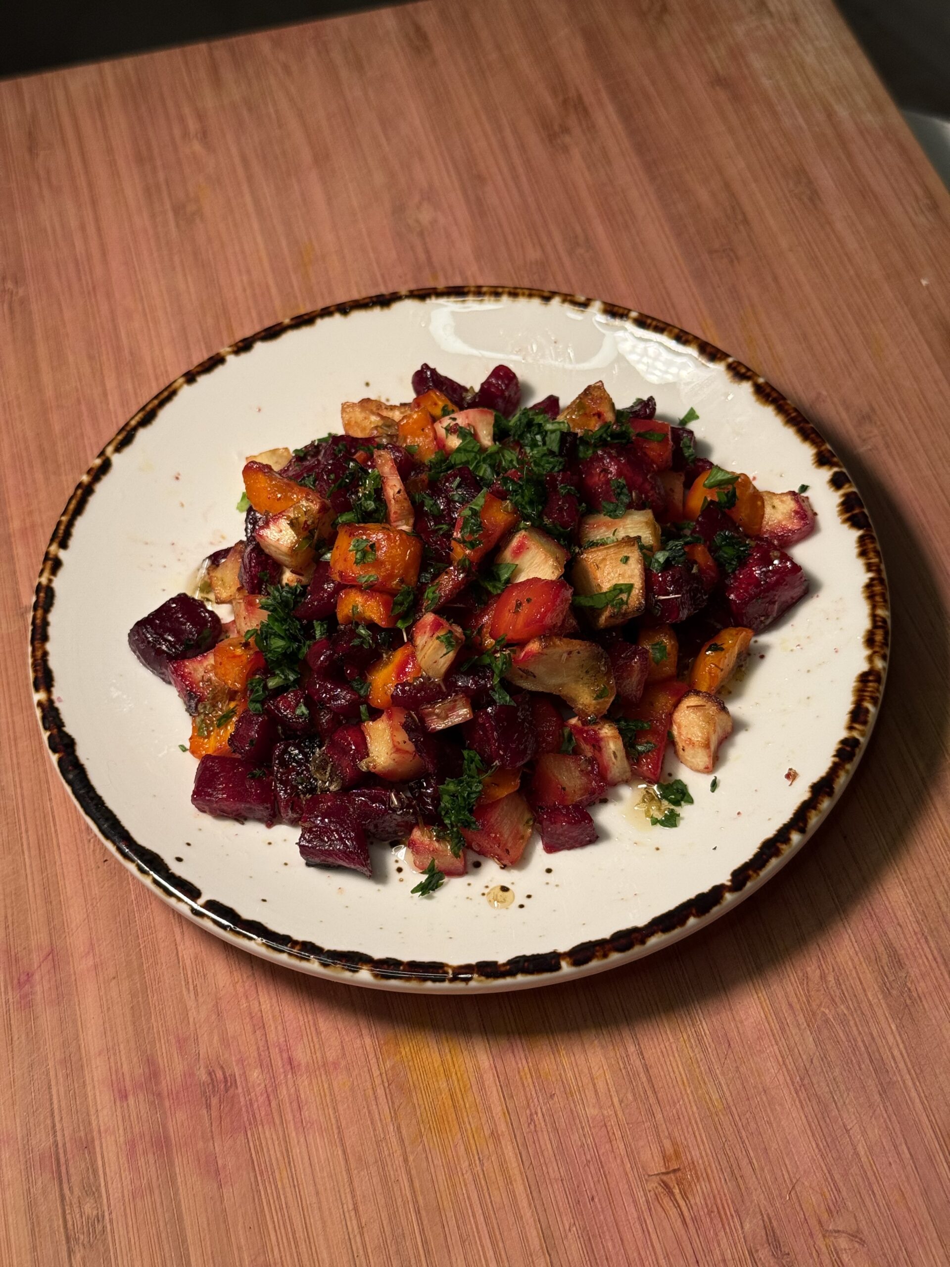 On a wooden cutting board, there is a white plate with cooked carrots, parsnips and beets garnished with green parsley. 