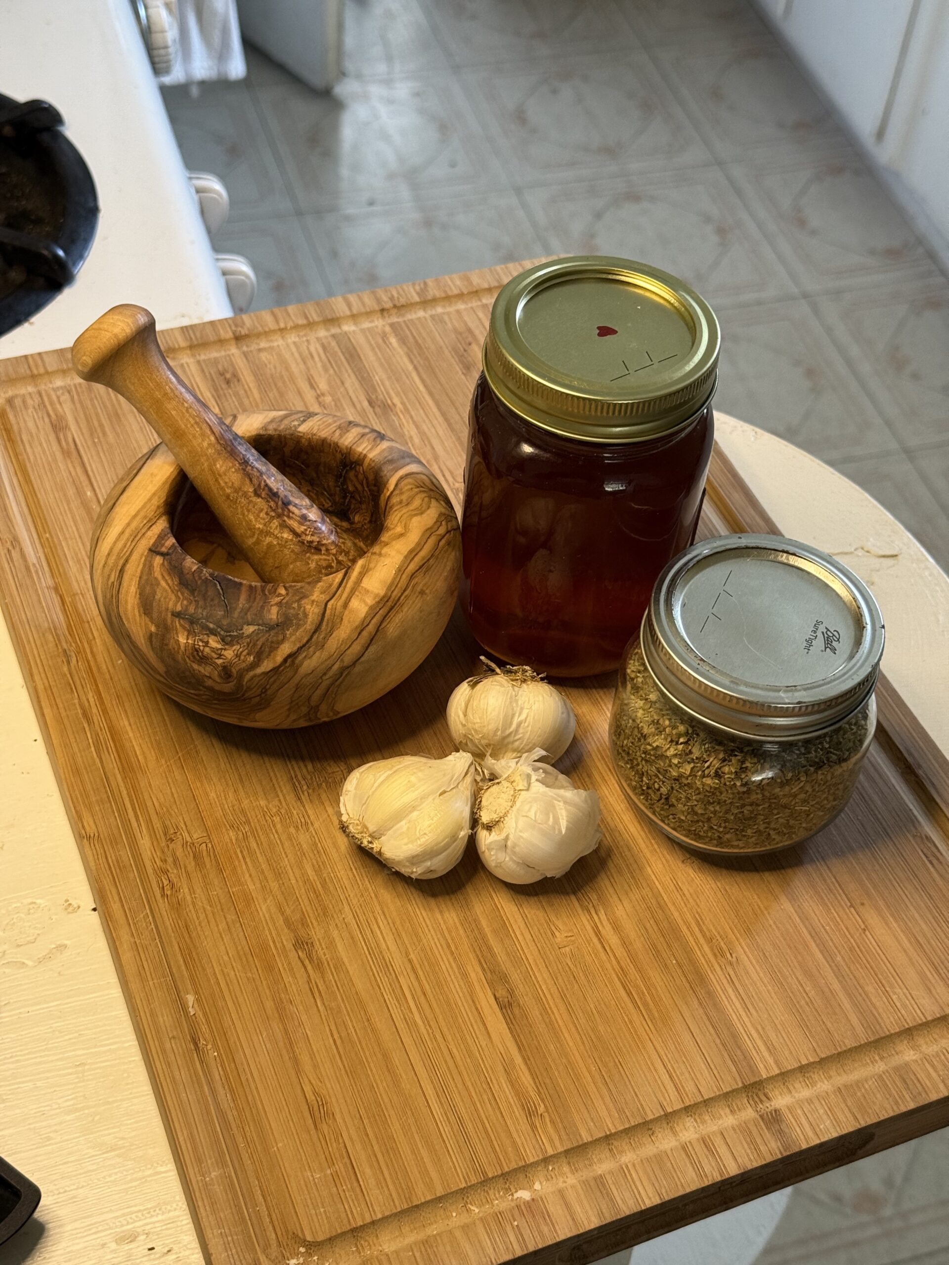 In a white kitchen, there is a wooden cutting board with a mortar and pestle next to a full jar of honey, three garlic heads, and a jar of dried oregano. 