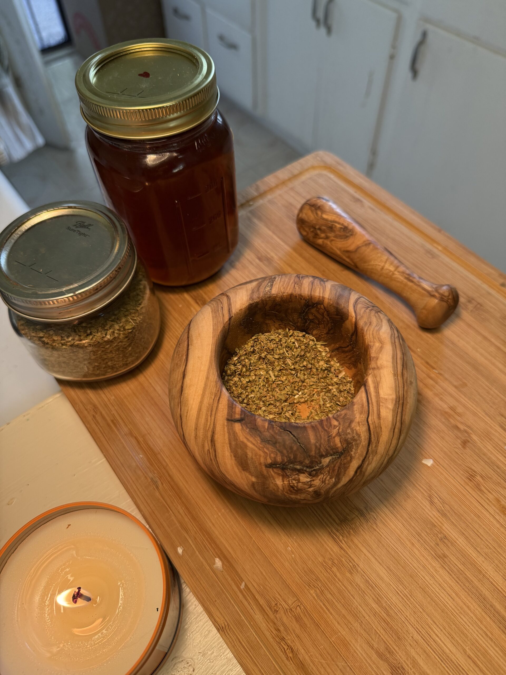In a white kitchen, there is a wooden cutting board with a lit candle sitting next to it on the white counter. On the cutting board, there is a wooden mortar and pestle filled with dried oregano with a jar of honey and a jar of oregano sitting behind. 