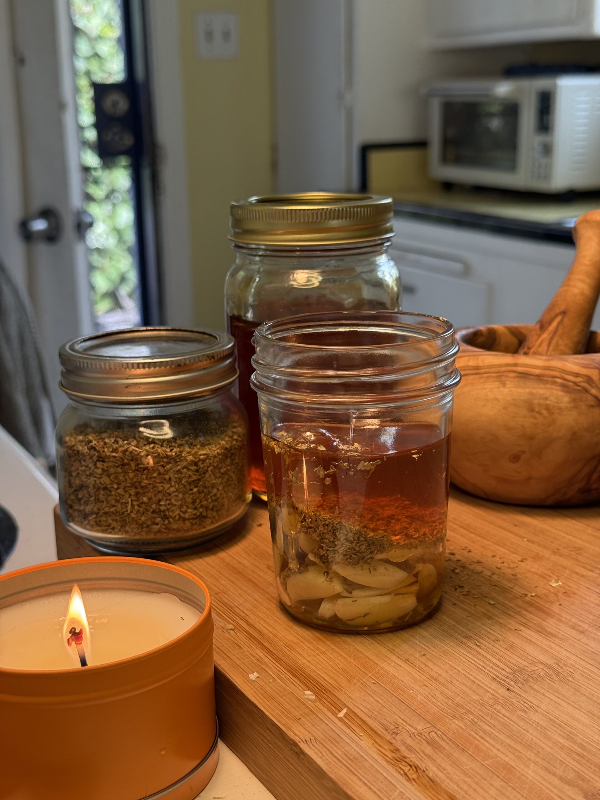 In a white kitchen, there is a open door revealing green leaves outside. In the kitchen, there is a lit candle in front of a wooden cutting board with a jar of dried oregano, a jar of honey, and a jar of sliced garlic, dried oregano and honey in one open jar with a wooden mortar and pestle on the side. 