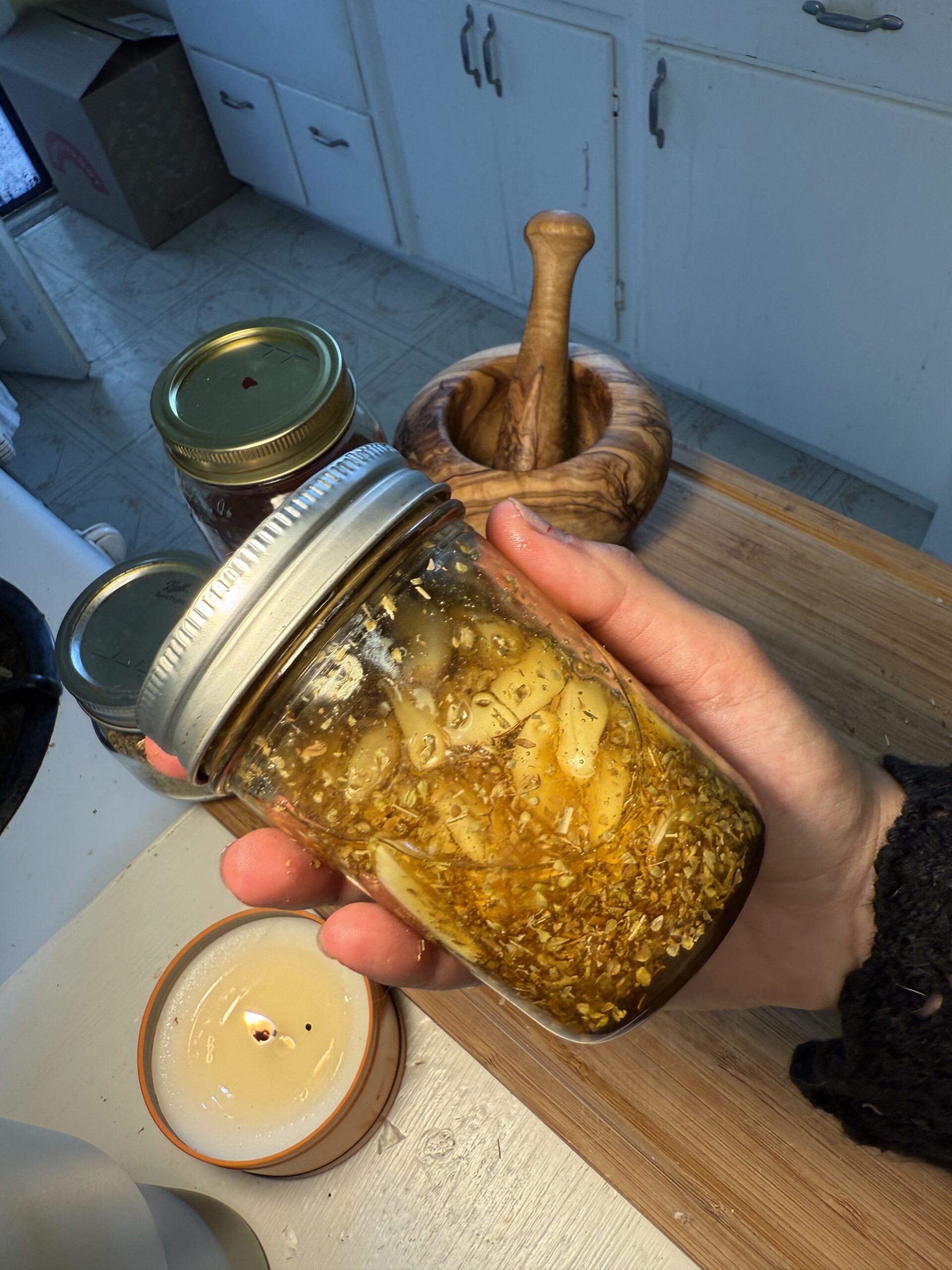 In a white kitchen, on a wooden cutting board, there is a wooden mortar and pestle, a jar of honey, a jar of oregano, and a white, lit, candle behind a hand holding a jar of honey, garlic, and oregano. 