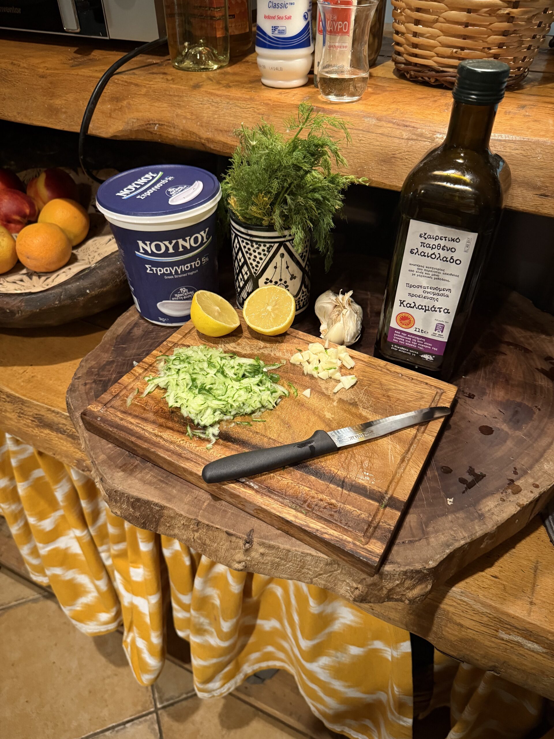 In a wooden kitchen, there is a cutting board with chopped green vegetables, garlic, and halved lemons. Behind the cutting board on the counter is a large bottle of olive oil, a bunch of garlic, a jar of green herbs, and a blue container of greek yogurt. 