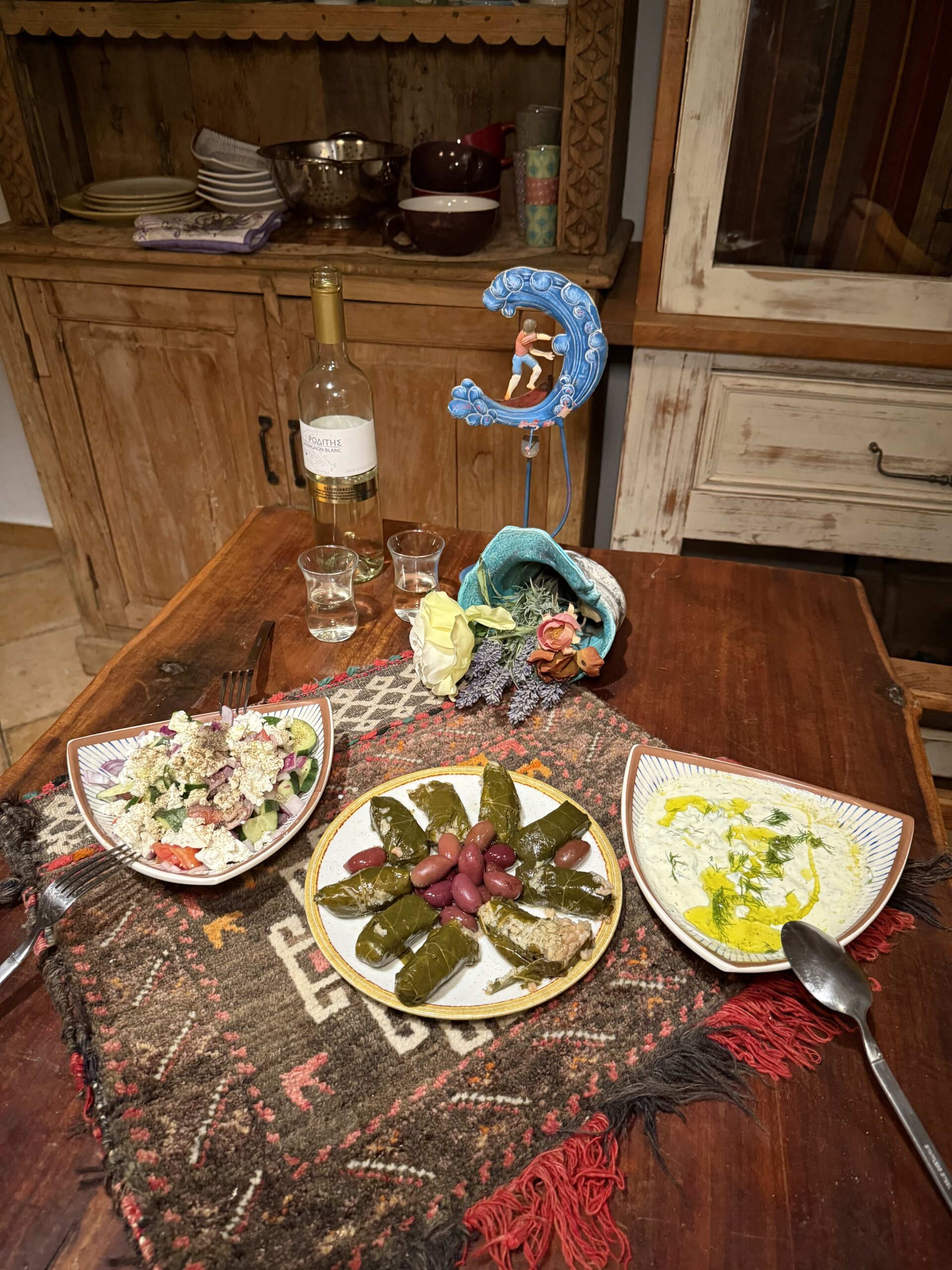 Rustic wooden table set with a Greek mezze platter featuring dolmades with Kalamata olives, fresh village salad, creamy tzatziki drizzled with olive oil, and a bottle of white wine, served on a woven table runner. 